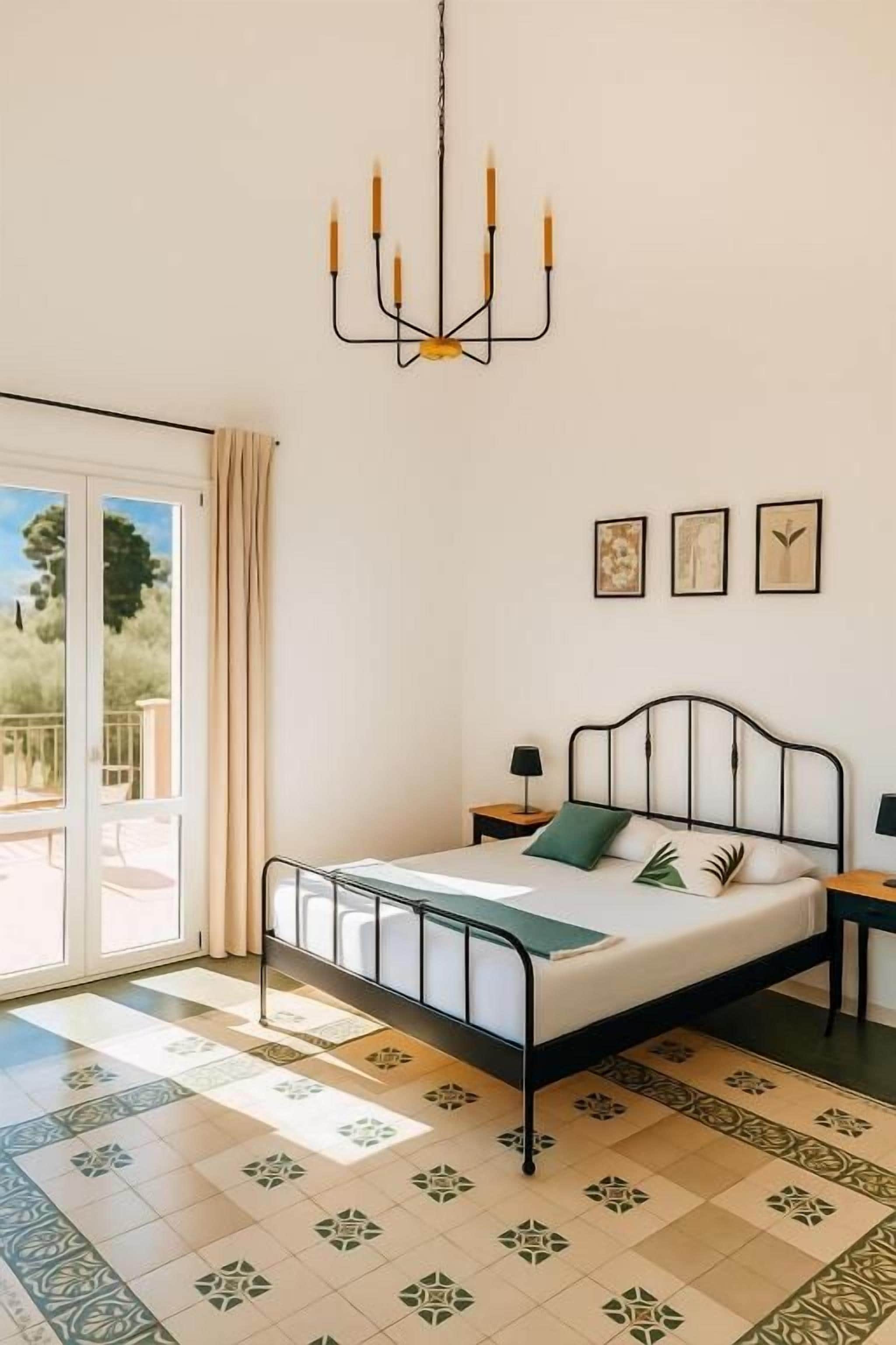 Un dormitorio con una cama, un candelabro, una ventana con cortinas y un piso estampado.