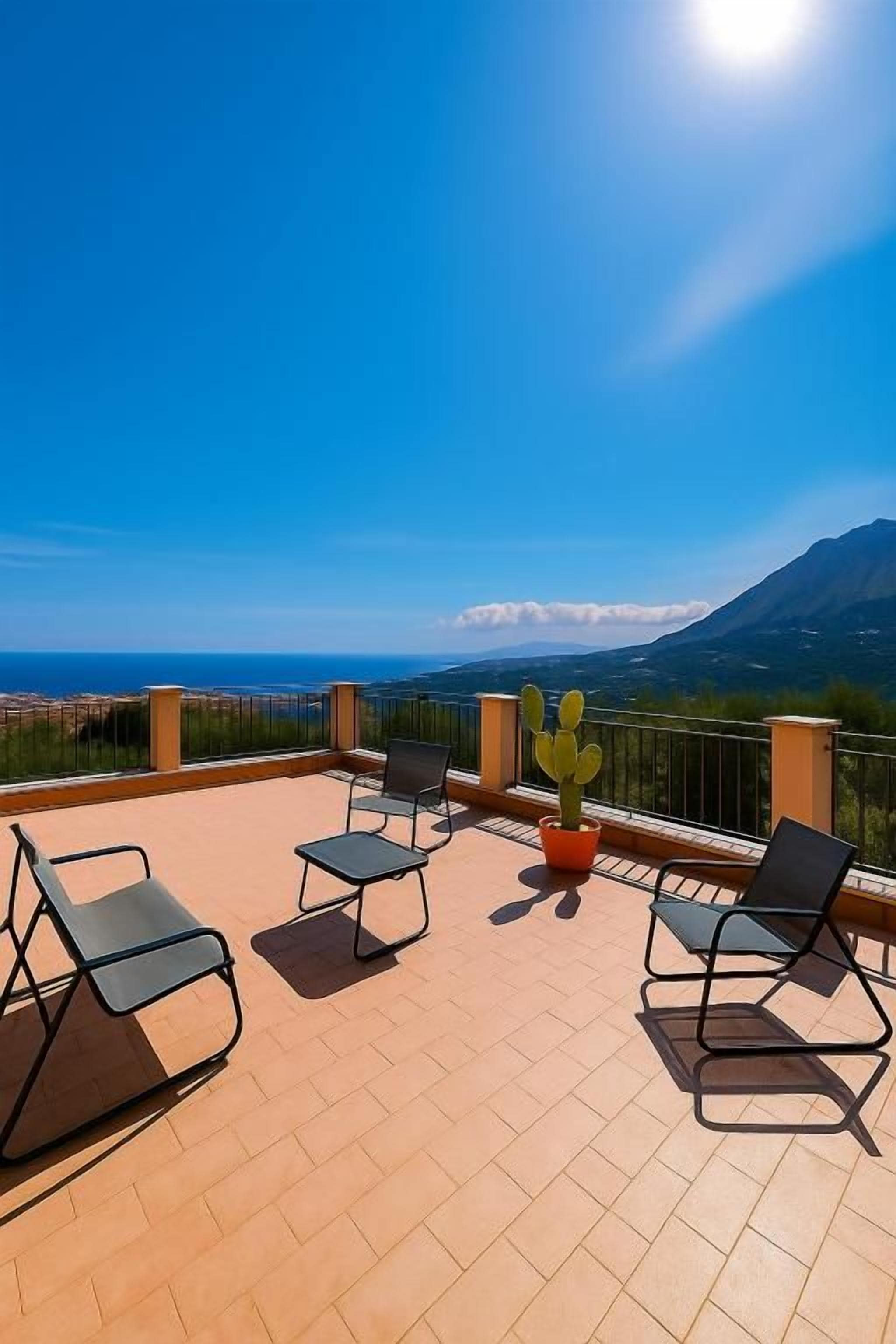 Una terraza con mobiliario exterior y una planta de cactus, con vistas a un paisaje costero con montañas.