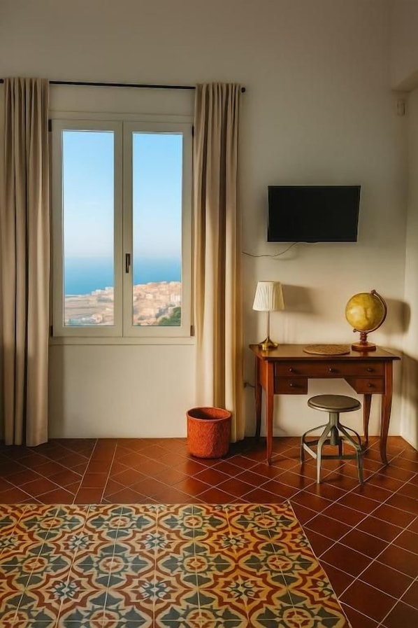 A room with a large window, a desk with a lamp, a globe, and a television on the wall.