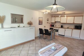 A modern kitchen with white cabinets, a dining area with a glass table, and a pendant light. - Holiday apartment "Horumersiel" (Wangerland)