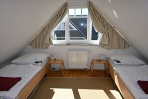 Een kleine, driekantige zolderkamer met twee eenpersoonsbedden, een radiator en een raam met gordijnen.