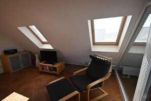 Ein kleiner Wohnbereich mit Fernseher, Büchern und einem Sessel. Sichtfenster und eine Heizung sind zu sehen.