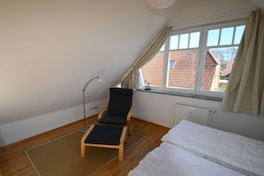 Ein Zimmer mit schrägen Wänden, einem Einzelbett, einem Sessel, einer Stehlampe und einem Fenster mit Vorhängen.