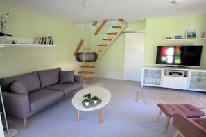 A modern living room with a grey sofa, a white coffee table with plants, a pink ottoman, a TV on a white cabinet, and a wooden spiral staircase. - Peter's holiday home (Ostseebad Ahrenshoop)