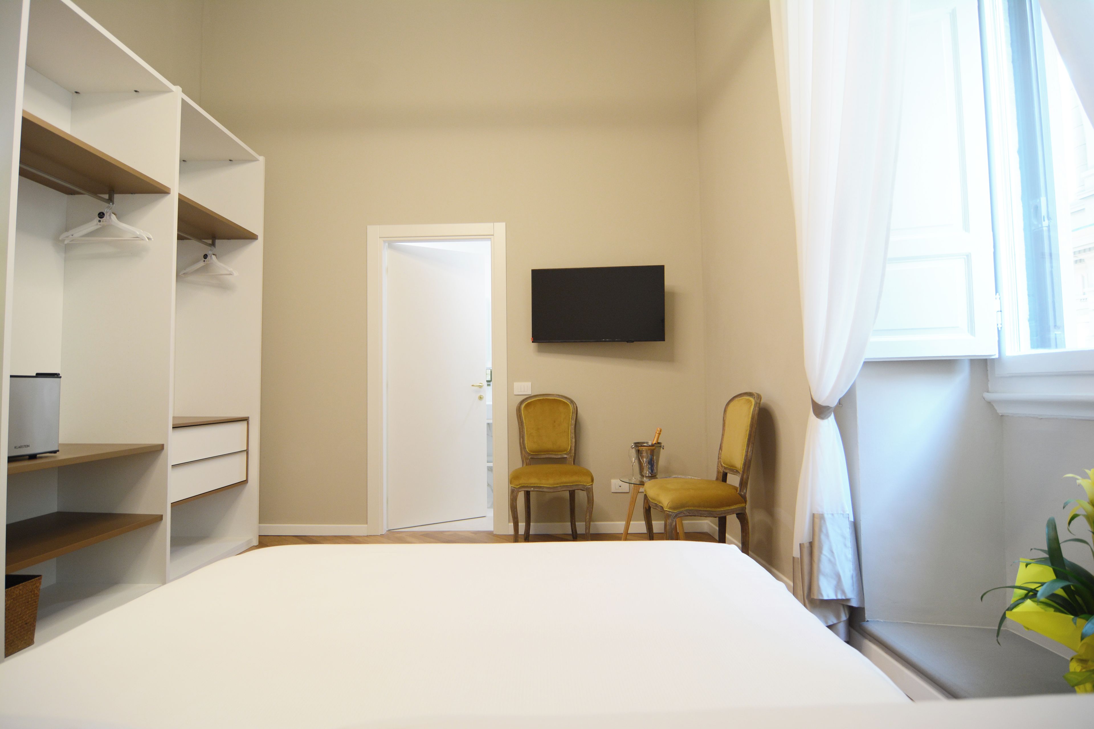 A bedroom with a bed, a wardrobe, two chairs, a television, and a window with curtains.
