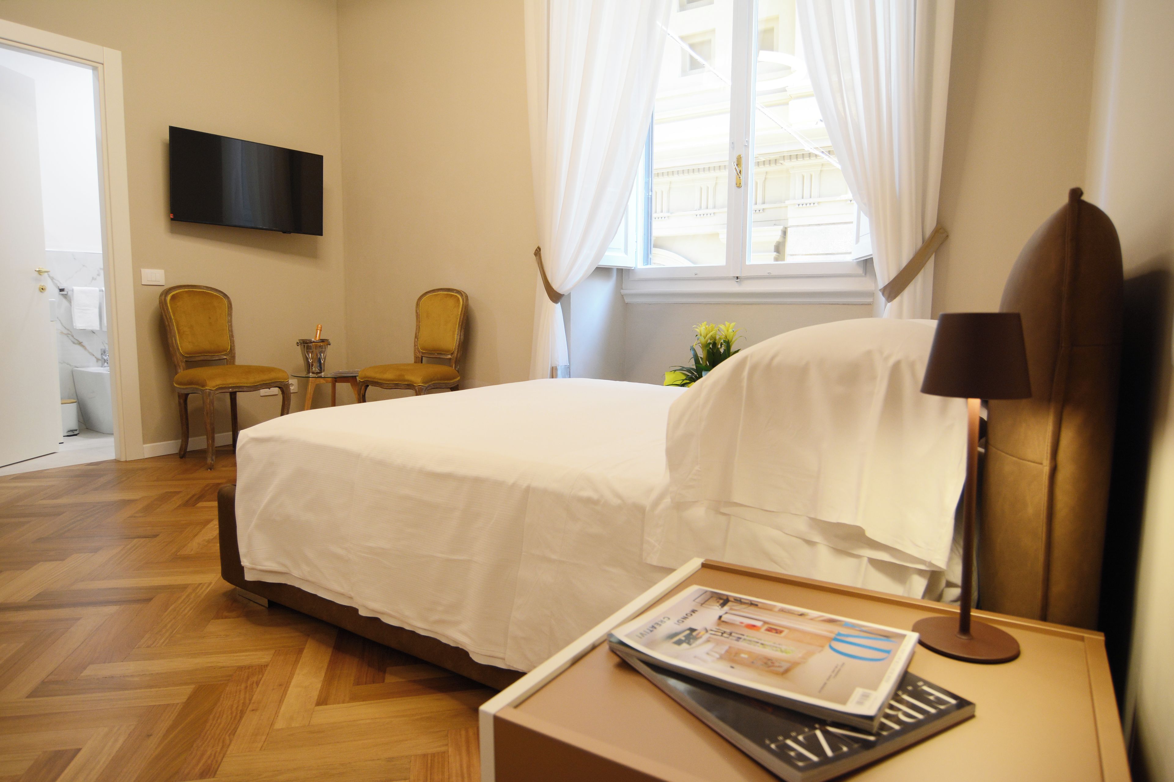 A hotel room with a bed, a chair, a table with a magazine, a lamp, and a TV on the wall.