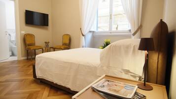 A hotel room with a bed, a chair, a table with a magazine, a lamp, and a TV on the wall.