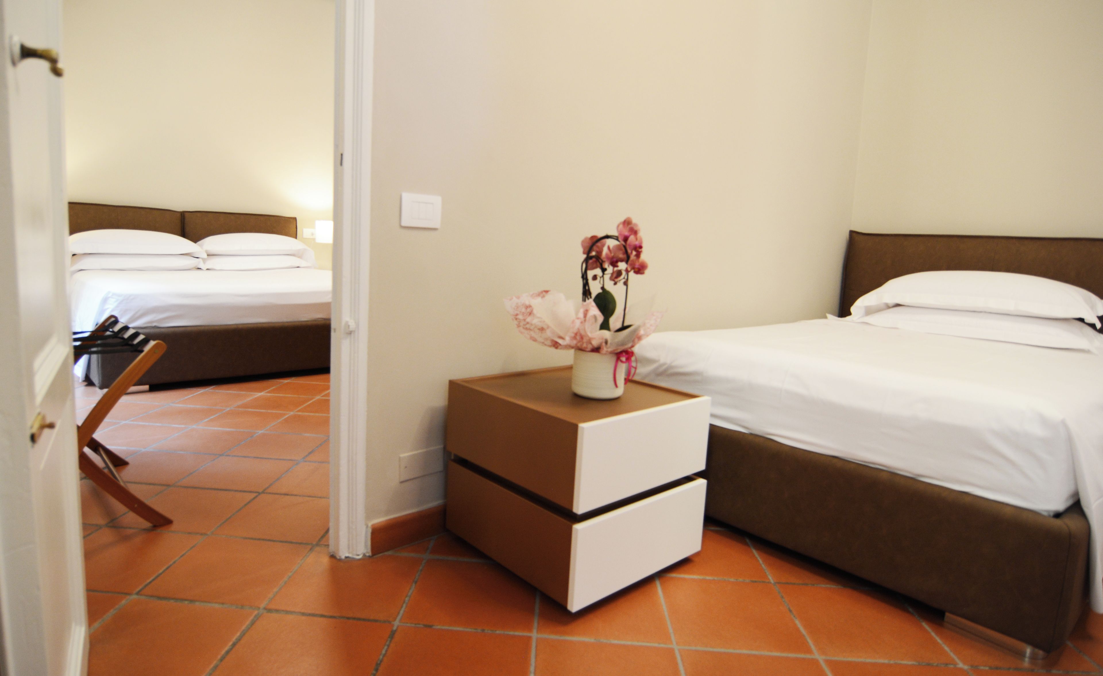 A hotel room with two beds, a nightstand with a vase of flowers, and a door leading to another room.