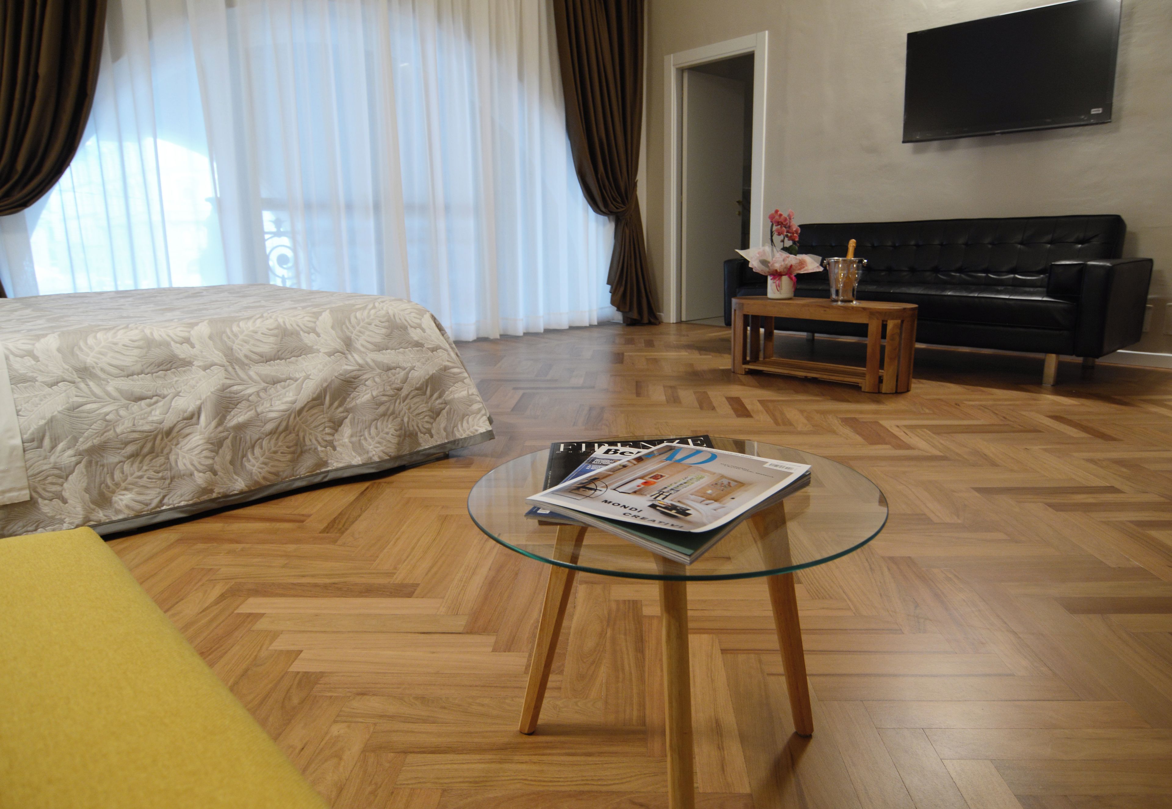 A hotel room with a bed, a glass coffee table with a magazine, a black sofa, and a wooden floor.