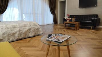 A hotel room with a bed, a glass coffee table with a magazine, a black sofa, and a wooden floor.