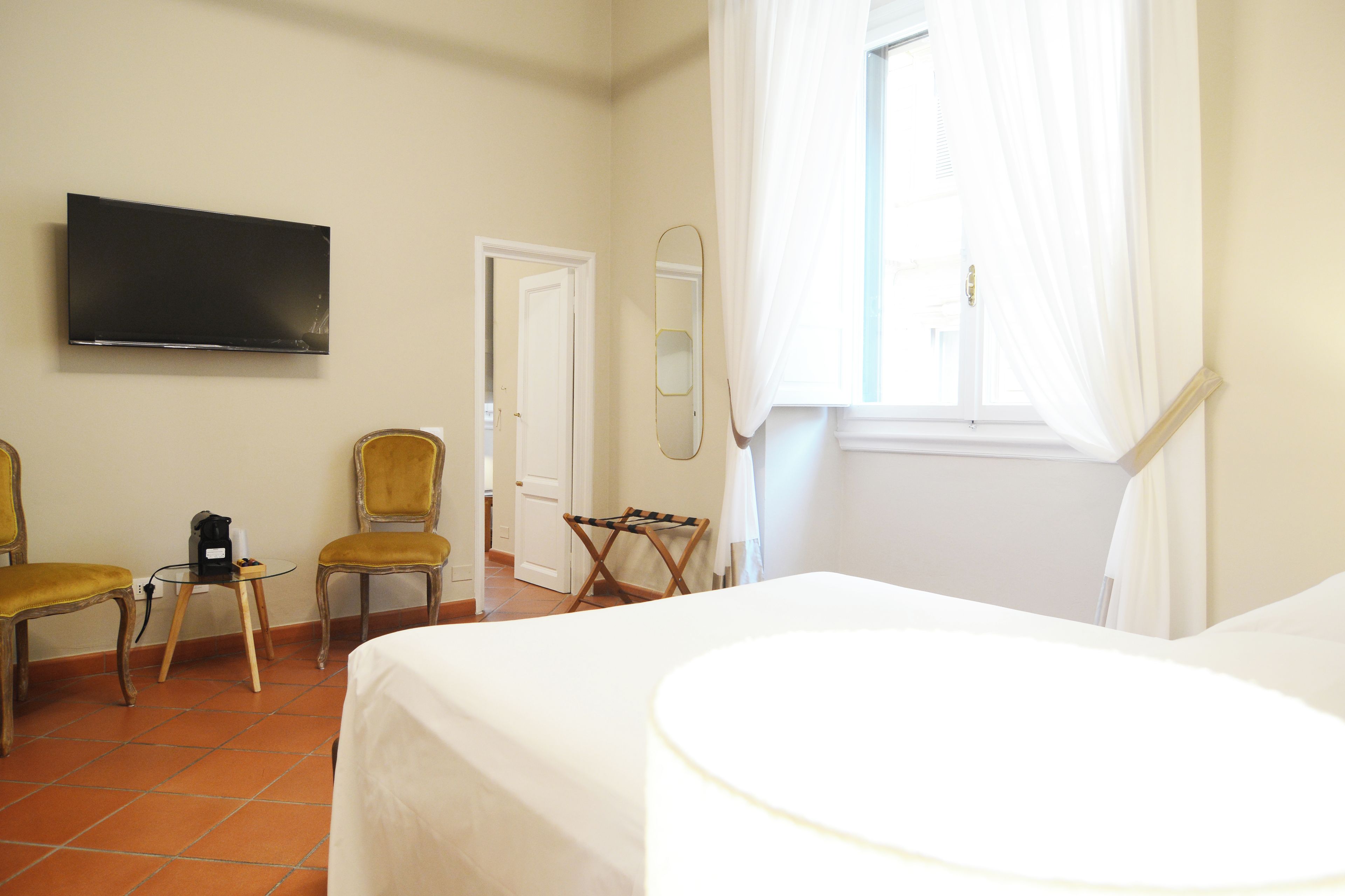 Ein Hotelzimmer mit einem Bett, einem an der Wand montierten Fernseher, einem Sessel, einem kleinen Tisch und einem Fenster mit Vorhängen.