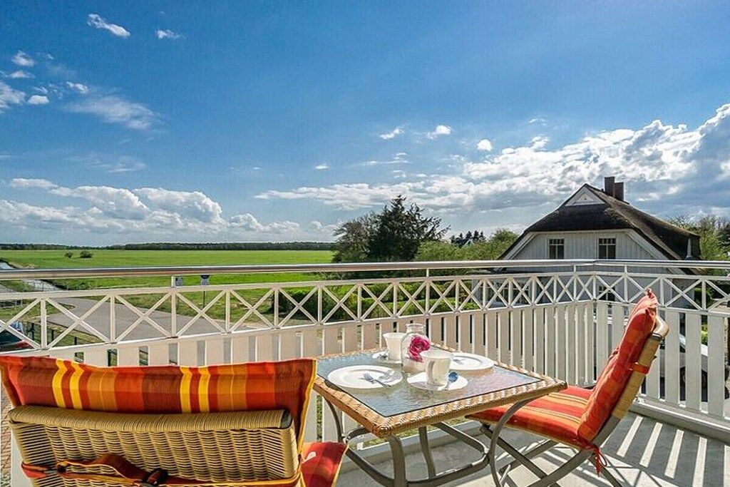 Un balcon avec des fauteuils en osier et une table, d’où l’on peut admirer un paysage verdoyant et une maison blanche.