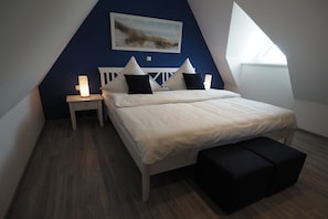 A bedroom with a sloped ceiling, a bed with white bedding, a nightstand with a lamp, and a painting on the wall. - Dream breeze "Dreaming under thatch" (Ostseebad Rerik)