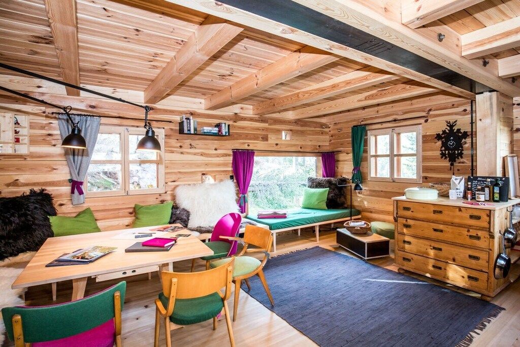 A cabin interior with a wooden ceiling, a dining table, chairs, a sofa, a dresser, and a window with curtains.