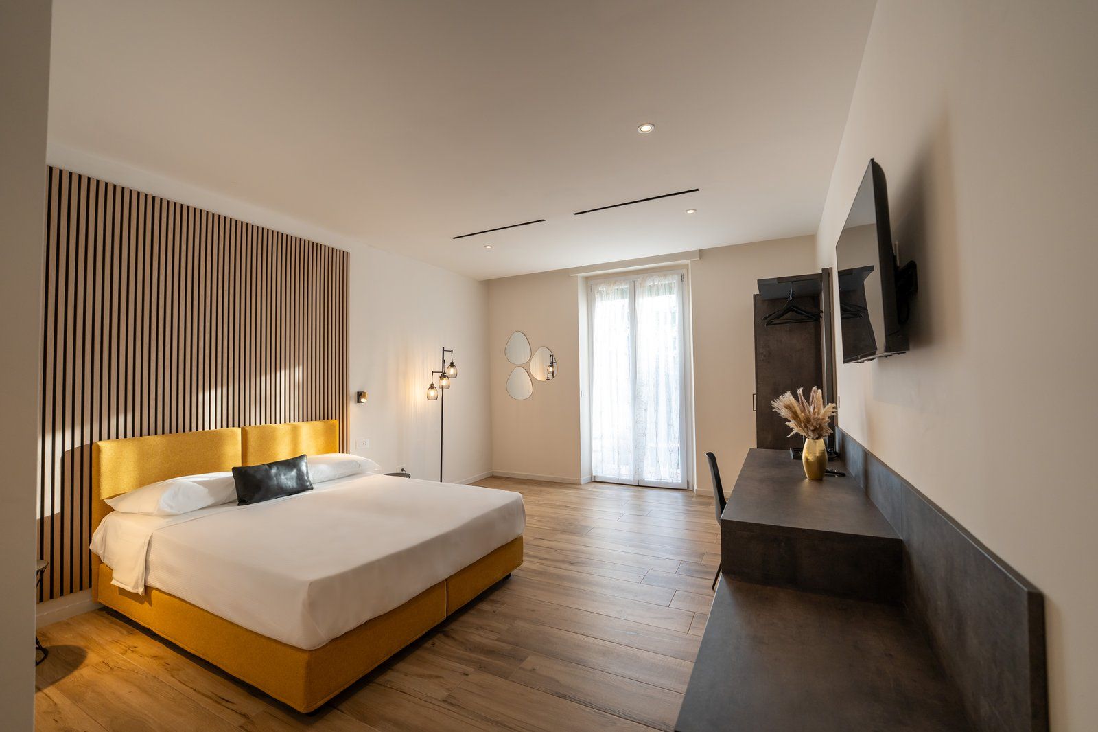 A modern bedroom with a large bed, a flat-screen TV mounted on the wall, and a sliding door leading to a balcony.