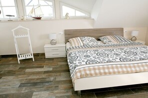 Ein Schlafzimmer mit einem Bett, Nachttischen, einer weißen Ablage mit Dekoration und einem Fenster mit einem Modell eines Segelboots.