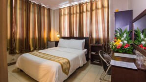 Quarto de hotel com cama, escrivaninha, cadeira, luminária e cortinas.