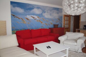 A living room with a red sofa, a white armchair, a white coffee table, and a large painting of a beach scene. - Karlshagen at the harbor (Karlshagen Usedom)