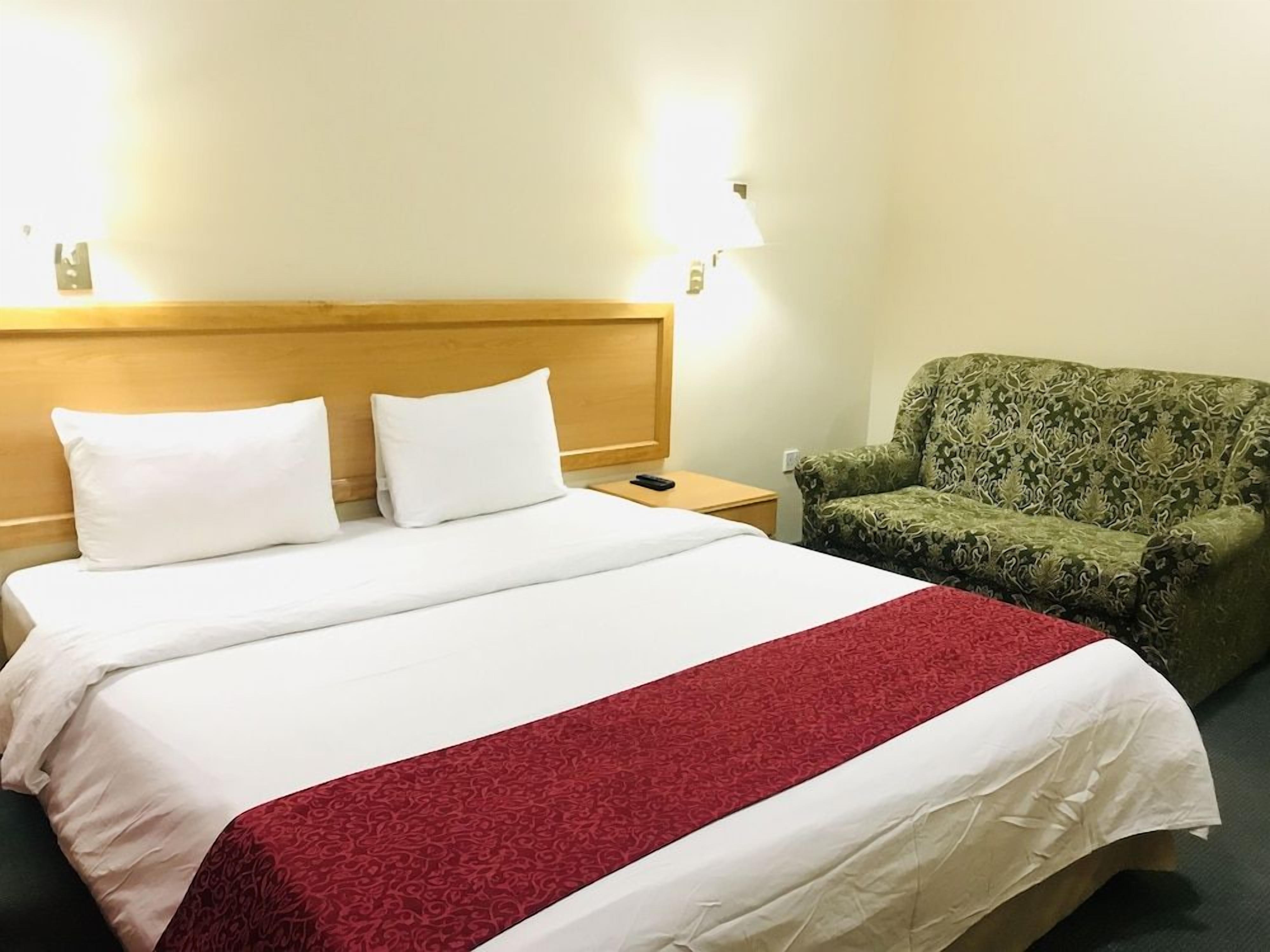 Habitación de hotel con cama, dos lámparas de noche, un sillón verde con estampado y una colcha blanca y roja.