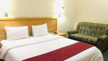 Habitación de hotel con cama, dos lámparas de noche, un sillón verde con estampado y una colcha blanca y roja.