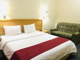 Habitación de hotel con cama, dos lámparas de noche, un sillón verde con estampado y una colcha blanca y roja.