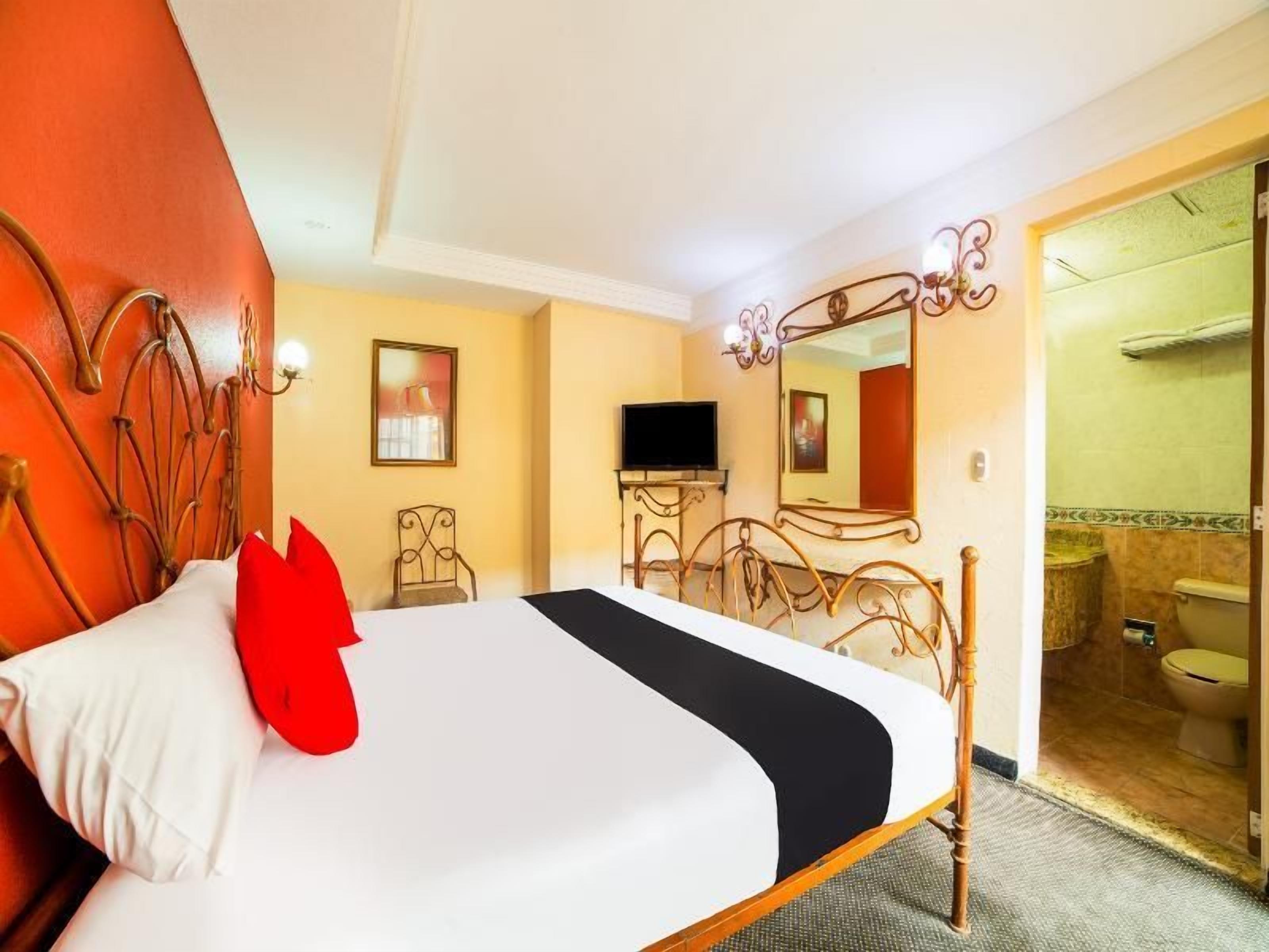 Habitación de hotel con cama, televisión, baño visible a través de una puerta abierta y espejo con luces decorativas.