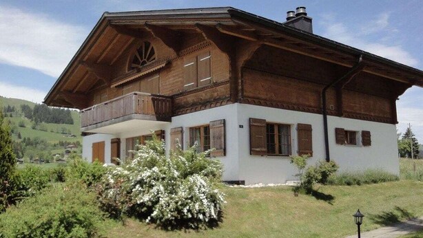 A traditional wooden chalet with a balcony, surrounded by lush greenery and a clear sky.