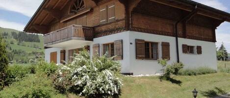 A traditional wooden chalet with a balcony, surrounded by lush greenery and a clear sky.