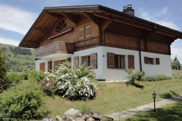 A traditional wooden chalet with a balcony, surrounded by lush greenery and a clear sky.