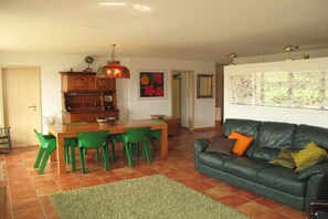 A living room with a green sofa, a dining table with chairs, a kitchen area with wooden cabinets, and a large wall art piece.