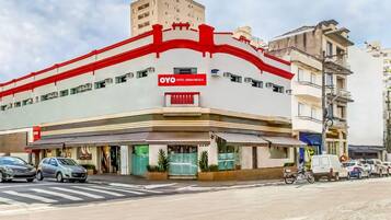 Hotel com fachada vermelha e branca marcante, entrada visível e estacionamento com carros e uma motocicleta.