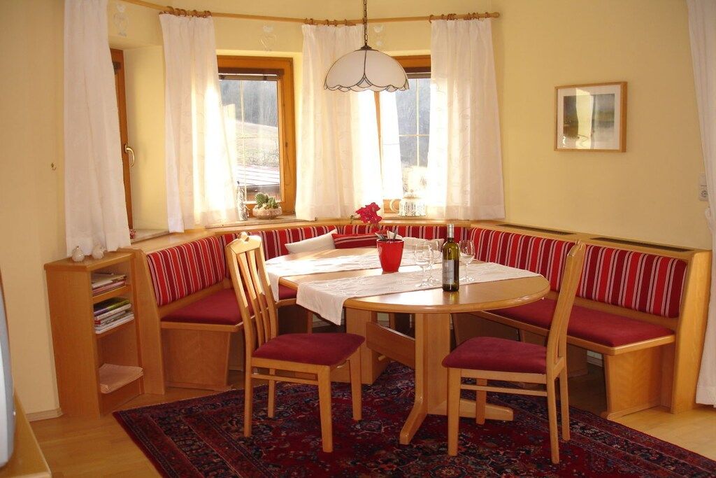 A dining area with a round table, wooden chairs, a bench with red cushions, a red vase, and a patterned rug.