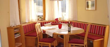 A dining area with a round table, wooden chairs, a bench with red cushions, a red vase, and a patterned rug.