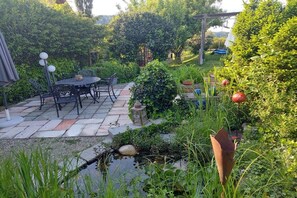 Une terrasse avec une table et des chaises, entourée d’une végétation luxuriante et agrémentée d’un petit élément décoratif aquatique.