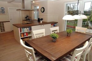 A dining area with a wooden table and white chairs, a kitchen with white cabinets, and a view through a glass door. - Holiday home with 2 bedrooms (Ostseebad Prerow)