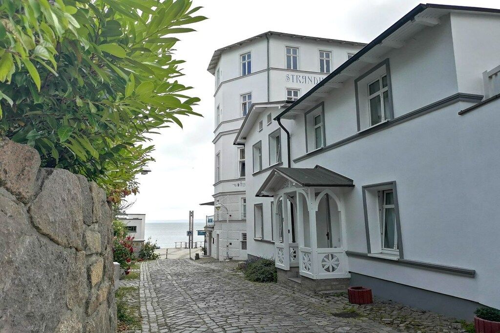 A cobblestone street leading to a row of white buildings with balconies, overlooking a body of water.