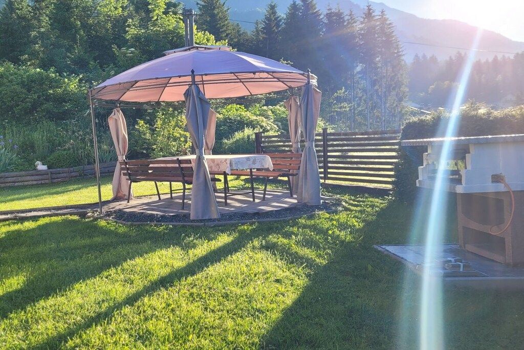 Ein Pavillon mit einer Bank, umgeben von einer Rasenfläche mit Holzzaun und Bäumen im Hintergrund.