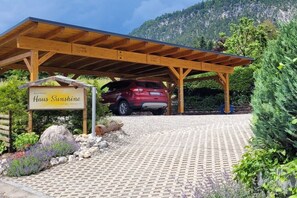 Un área de estacionamiento techada con una camioneta SUV roja, un letrero que dice 'Haus Sunshine' y montañas al fondo.