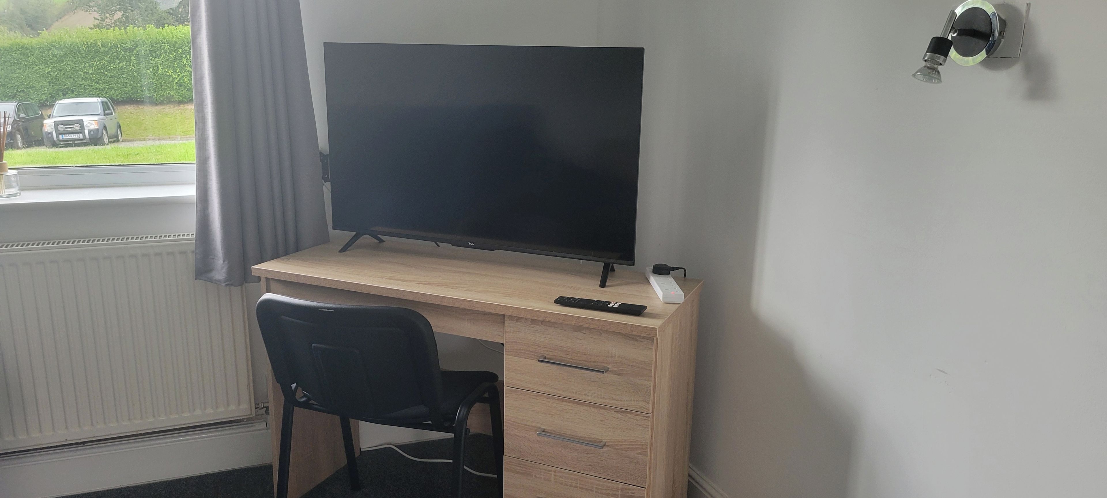 A room with a wooden desk, a black chair, a flat-screen TV, and a window with a view of parked cars and greenery outside.