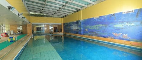 Une piscine intérieure avec une fresque murale en mosaïque, une terrasse en bois et un plafond doté de lucarnes.