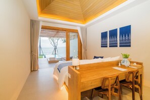A hotel room with a wooden bed, a desk, and chairs, a view of the sea, and three wall-mounted paintings.
