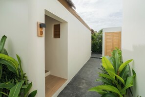 Un pasillo estrecho con una puerta de madera al final, una ventana pequeña y macetas a ambos lados.