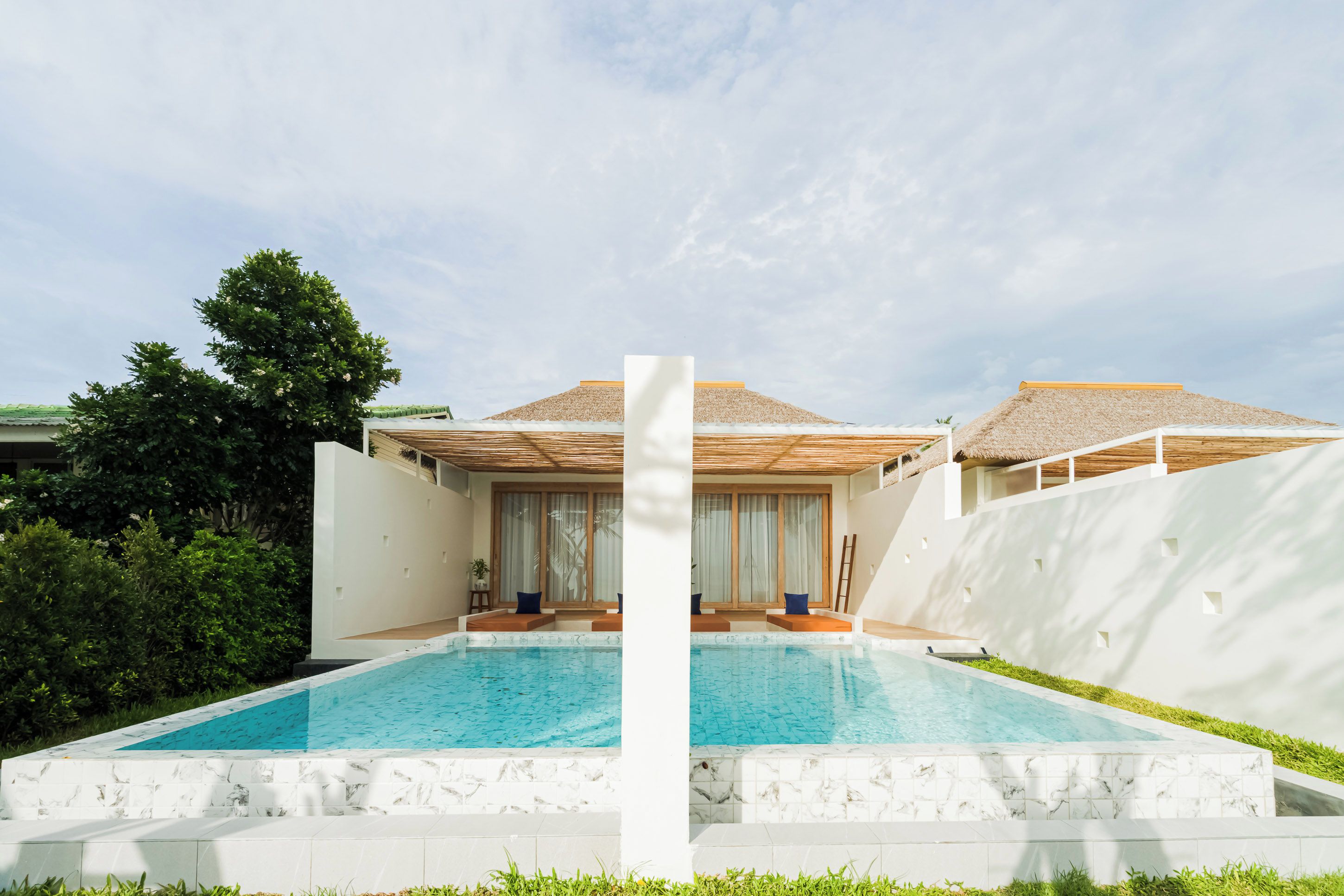 Un área moderna junto a la piscina con una pileta cristalina, paredes blancas y una estructura con techo de paja.