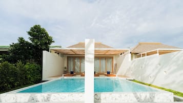 Un área moderna junto a la piscina con una pileta cristalina, paredes blancas y una estructura con techo de paja.