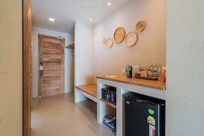 A hotel room with a wooden door, a small kitchenette with a refrigerator, a coffee machine, and a shelf with toiletries.