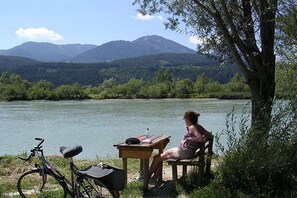 Eine Person sitzt an einem Tisch am Flussufer, ein Fahrrad und ein Rucksack stehen in der Nähe.