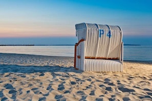 A beach chair with a number '4' on it, positioned on a sandy beach with a calm sea and a pier in the background. - "Beach Promenade" - Haus Strelasund (Ostseebad Binz)