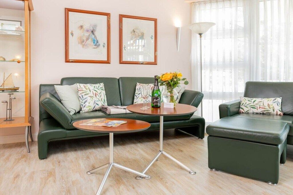 Un salon moderne avec un canapé vert, une table basse ronde et un pouf assorti. Deux œuvres d’art encadrées ornent le mur, ainsi qu’une lampe sur pied et une fenêtre agrémentée de rideaux légers.