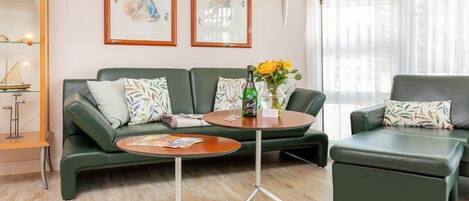 Un salon moderne avec un canapé vert, une table basse ronde et un pouf assorti. Deux œuvres d’art encadrées ornent le mur, ainsi qu’une lampe sur pied et une fenêtre agrémentée de rideaux légers.