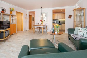 A modern living room with a green sofa, a dining area with a table and chairs, a kitchen in the background, and a television on a wooden cabinet. - "Beach Promenade" - Haus Strelasund (Ostseebad Binz)